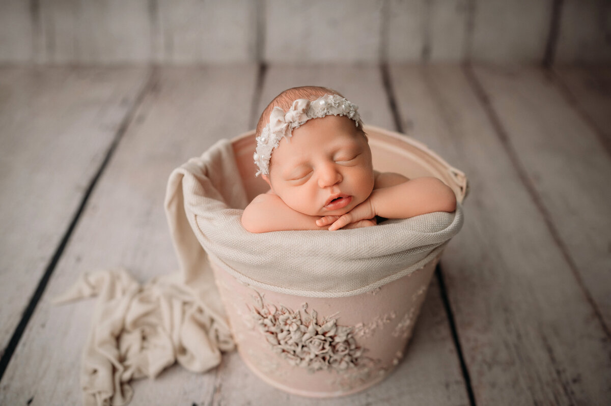 newborn baby girl wearing cream headband posed in bucket with chin on hands on cream wooden plank backdrop