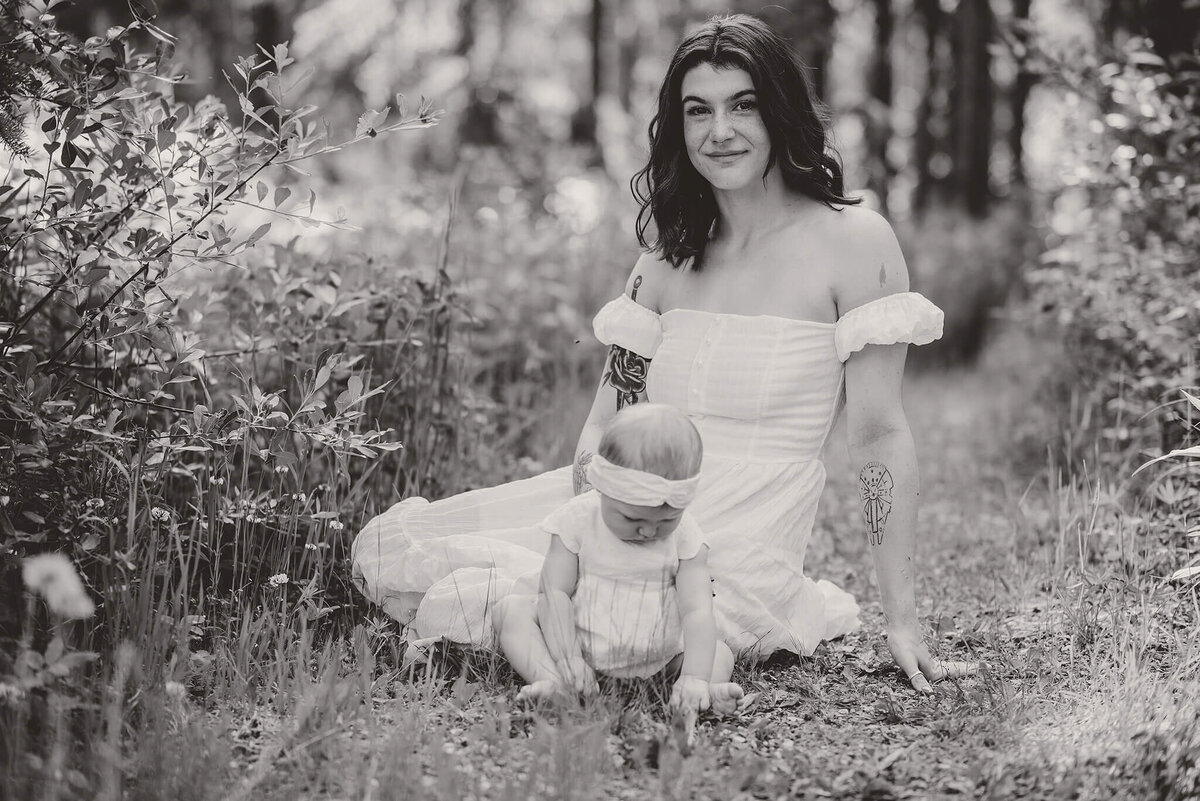 A black and white image of a mother and daughter sitting on the ground amongst tall grass. Mom smiles at the camera while baby daughter plays on the ground. Taken by Brittany Danielle