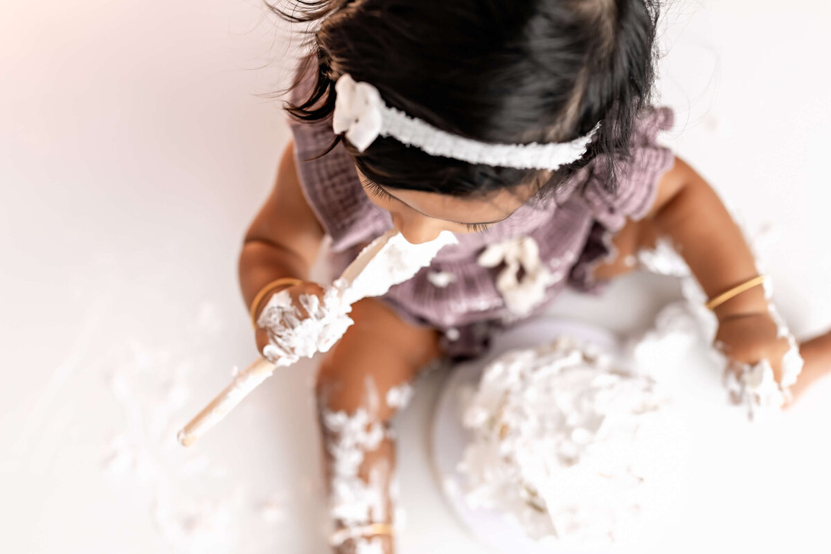 A young child with a headband sits on the floor, eating and playing with a cake covered in white frosting. The cake and the child’s hands are messy with frosting. The child is wearing a purple outfit.