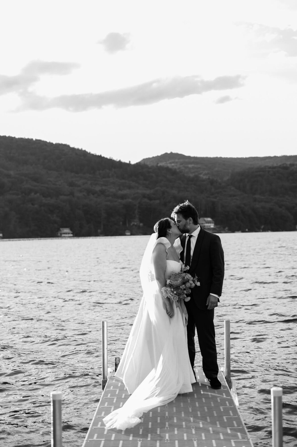 Black and white photo of a bride and groom kissing on a dock with the lake and Vermont mountains in the background.