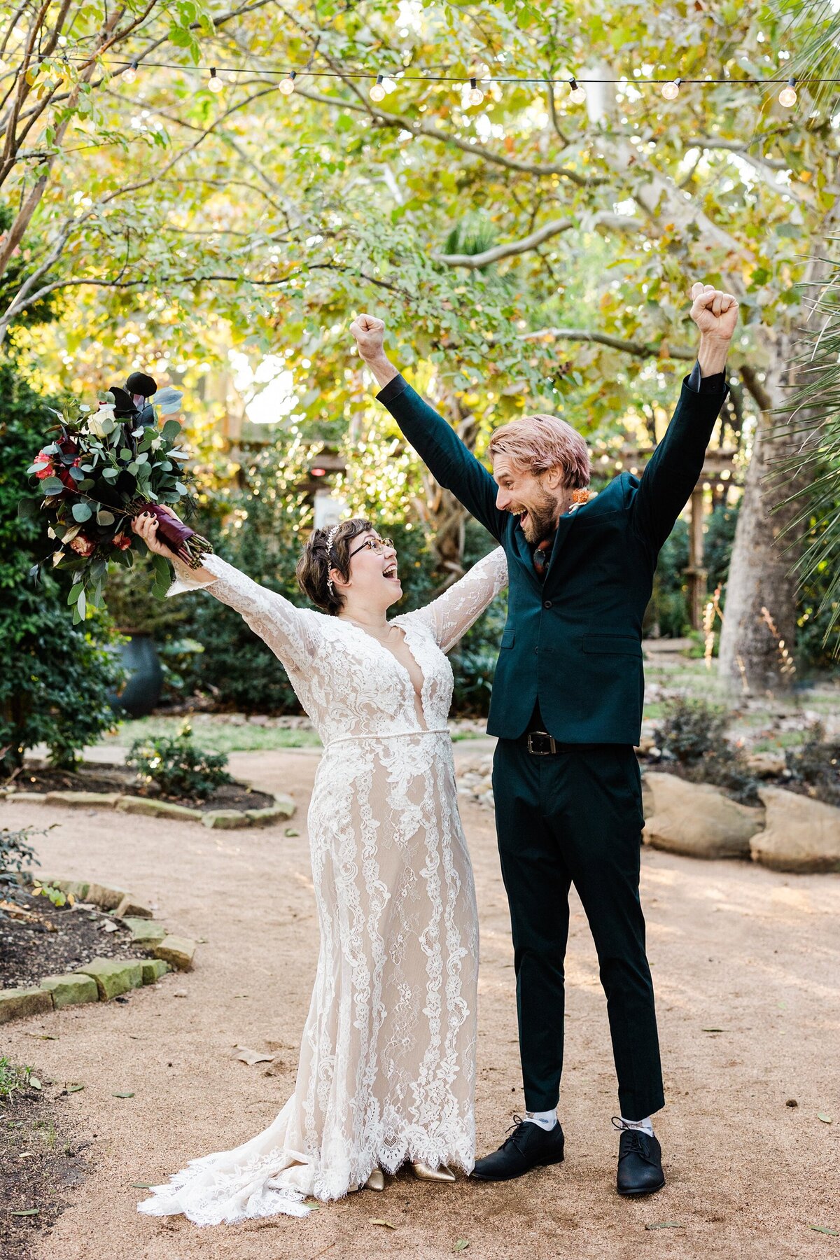 A fun, playful portrait of a bride and groom cheering triumphantly after their wedding ceremony in Dallas, Texas. The bride is on the left and is wearing a long sleeve, intricate, white dress with a floral headband and a large bouquet. The groom is wearing a dark suit with a boutonniere. They both hold their arms up in celebration while being framed by many trees and other natural elements.