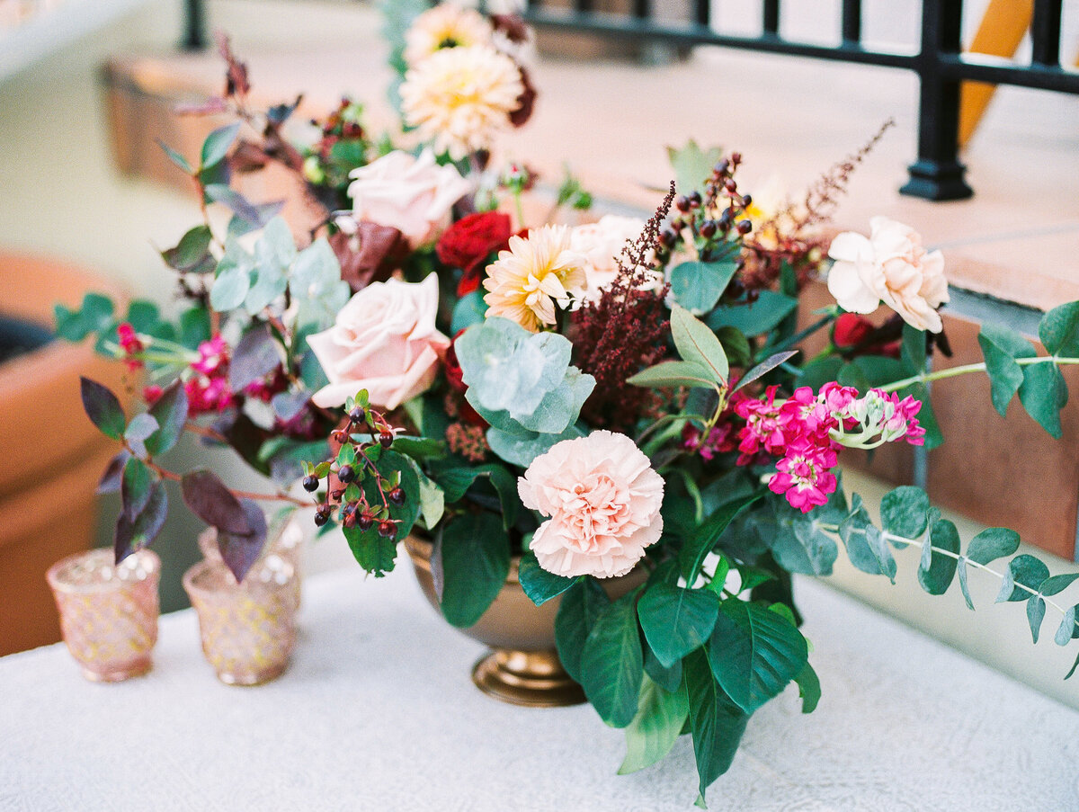 Romantic wedding centerpiece at the iconic Kimpton Canary Hotel rooftop in Santa Barbara. Organic garden-style arrangement featuring blush carnations, burgundy astilbe, peach dahlias, and vibrant pink bougainvillea in a gold compote vase. Lush tropical monstera leaves and eucalyptus add Santa Barbara coastal flair. Mediterranean-inspired wedding decor with vintage pink and gold votives. Luxury Southern California rooftop wedding florals with ocean and mountain views.