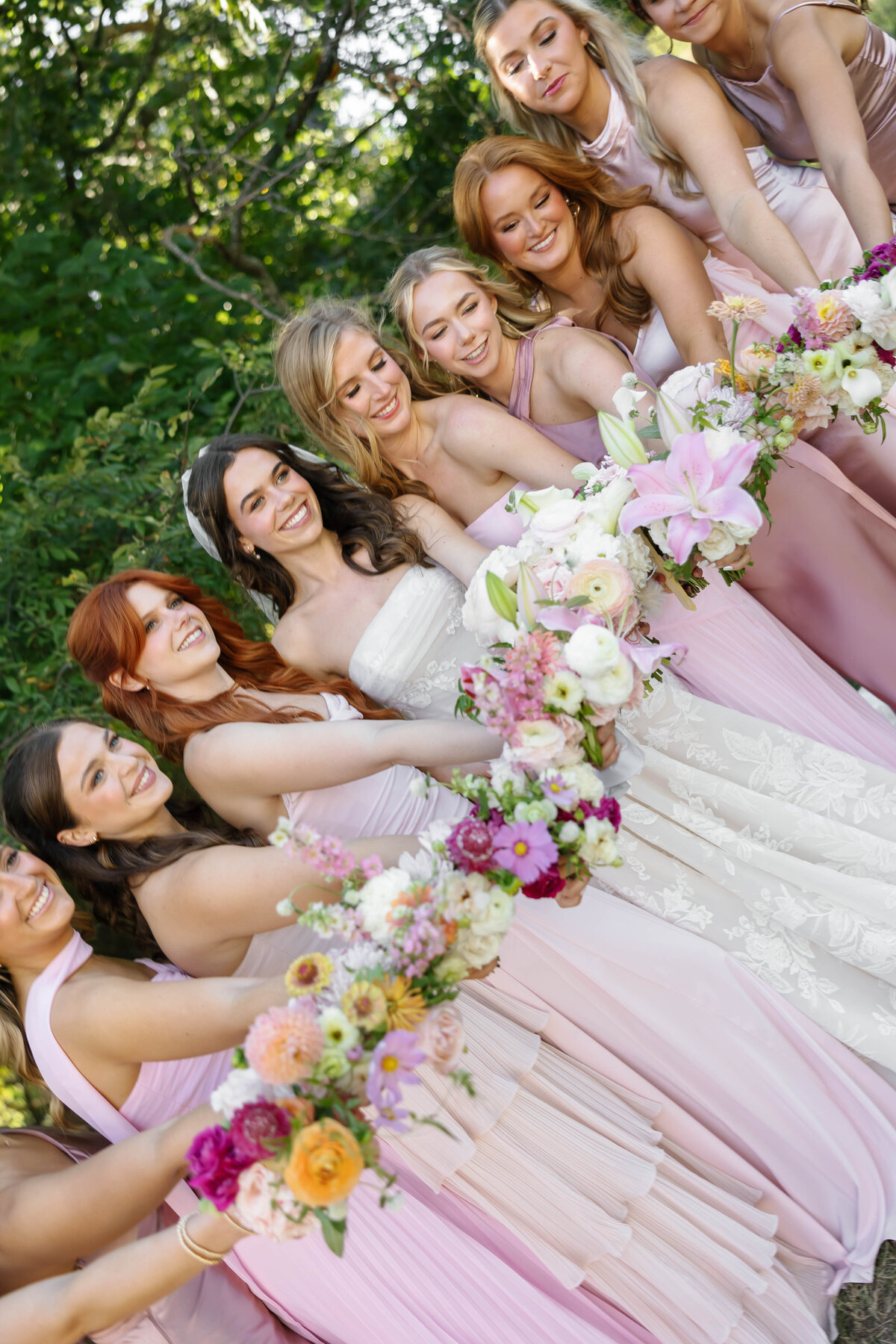 Bridesmaids and bride standing together holding colorful floral bouquets, smiling and leaning in during outdoor wedding portraits in natural greenery, soft pink dresses and vibrant flowers.