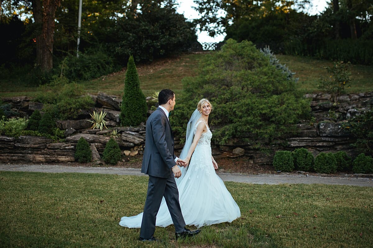 The bride, wearing a long sleeveless lace wedding dress and veil takes the groom, wearing a charcoal gray suit, by the hand as they walk in the garden at Cheekwood