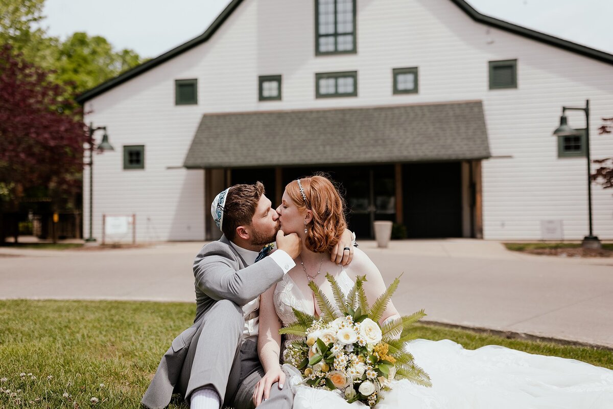 The groom, wearing a grey suit and white kippot kisses the redheaded bride. The bride, wearing a white lace wedding gown with a plunging neckline holds a large bouquet of white, yellow and blush wild flowers with fern and greenery as they sit on the ground in front of a large white barn with green trim at Loveless Cafe in Nashville, TN