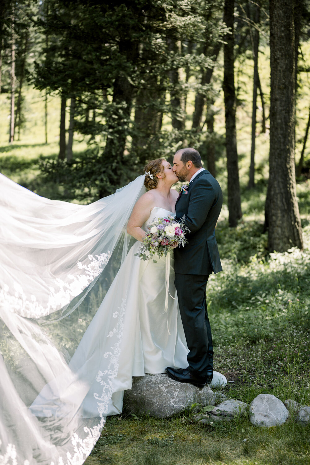 Bride and groom sharing a kiss with the bride’s cathedral veil  flowing in the wind during their Montana summer wedding. 