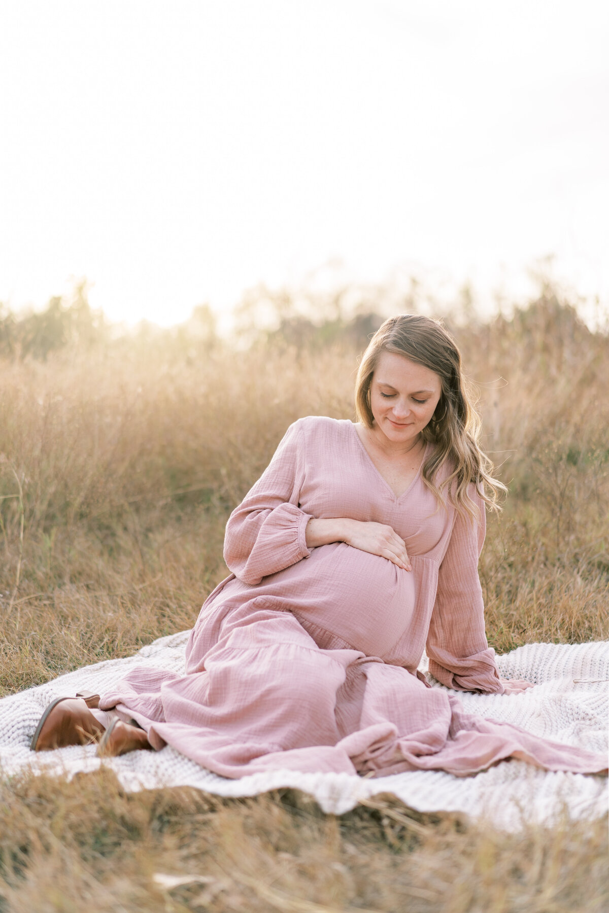 Portrait of a pregnant woman wearing a blush dress sitting atop a waffle blanket in the country during golden hour.