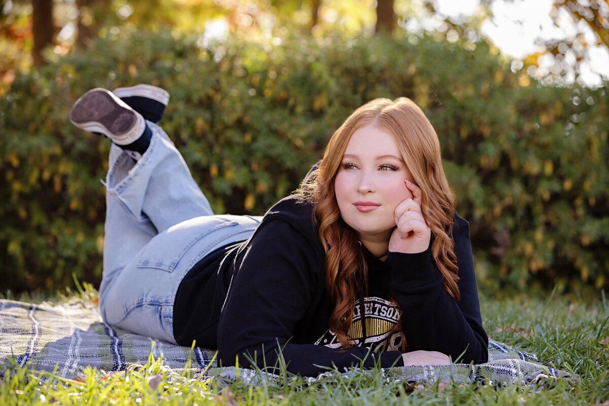 High school senior in black SENIORS hoodie laying on a blanket in grassy park with a sweet, reflective smile