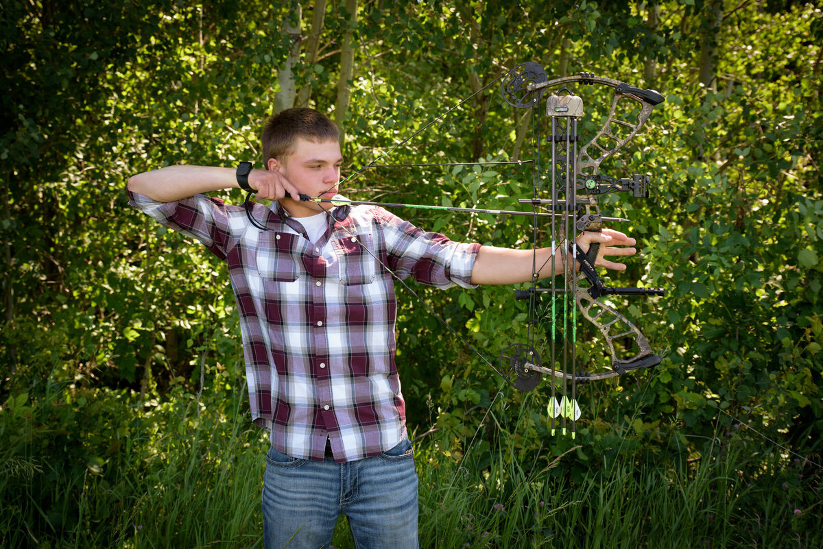 Luxemburg Casco high school senior boy wearing a maroon, black and white plaid button down shirt and jeans holding a hunting bow in farm field at his home near Green Bay, Wisconsin.