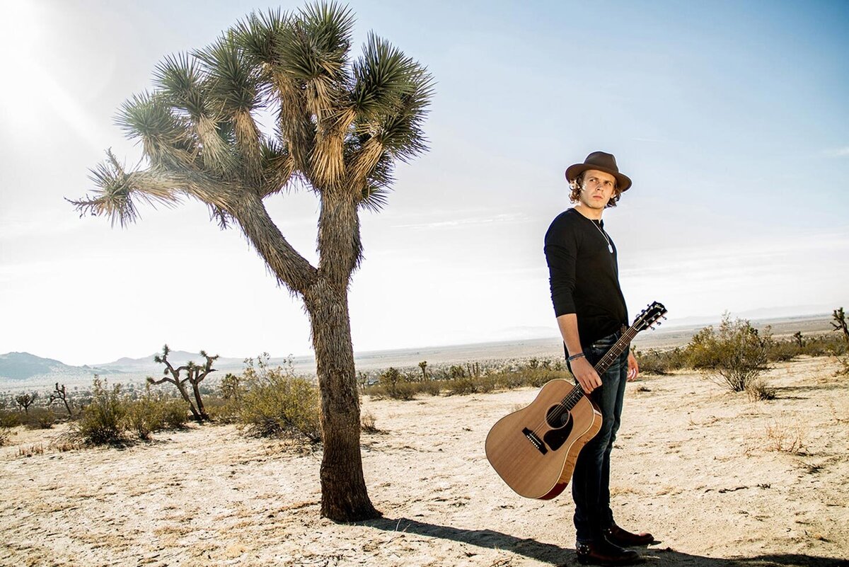 Musician Photo Cole Bradley standing in desert next to joshua tree holding acoustic guitar by neck base Just Play Something Gallery