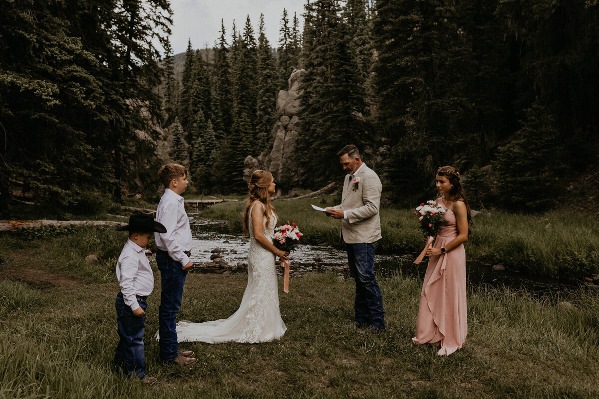 a bride and groom standing together during their intimate wedding ceremony in front of a creek with their kids