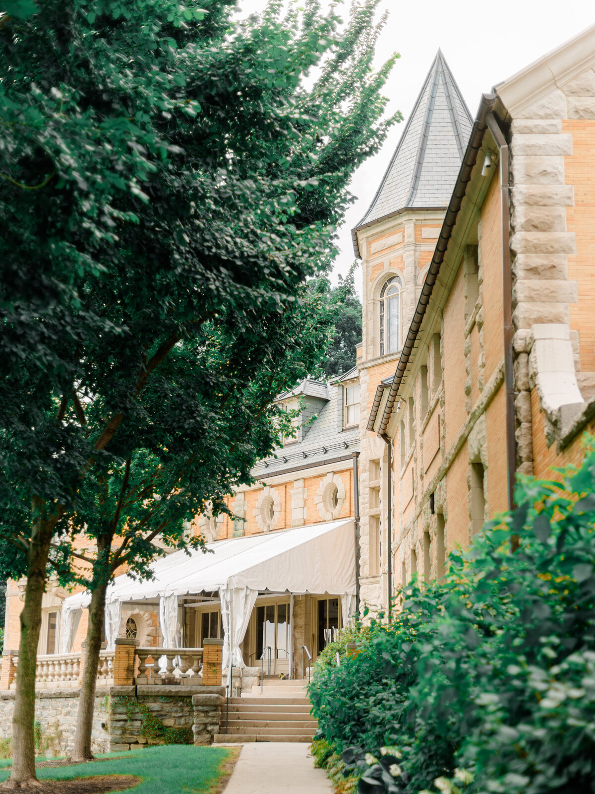 A view of the historic Cairnwood Estate, showcasing its French-inspired architecture, stone detailing, and tented terrace framed by lush greenery. This romantic setting, perfect for elegant East Coast weddings, is captured by Elizabeth Kane Photography, a Luxury Philadelphia Wedding Photographer.