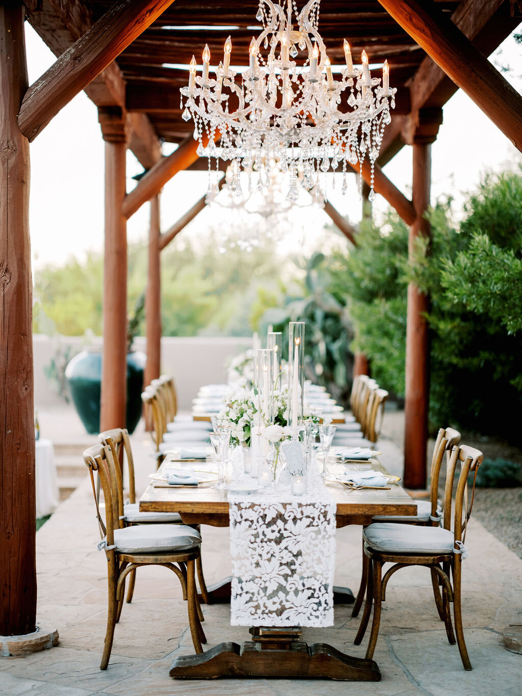 A dreamy outdoor reception table setup featuring a rustic wooden table, crystal chandeliers, and romantic candlelit decor.