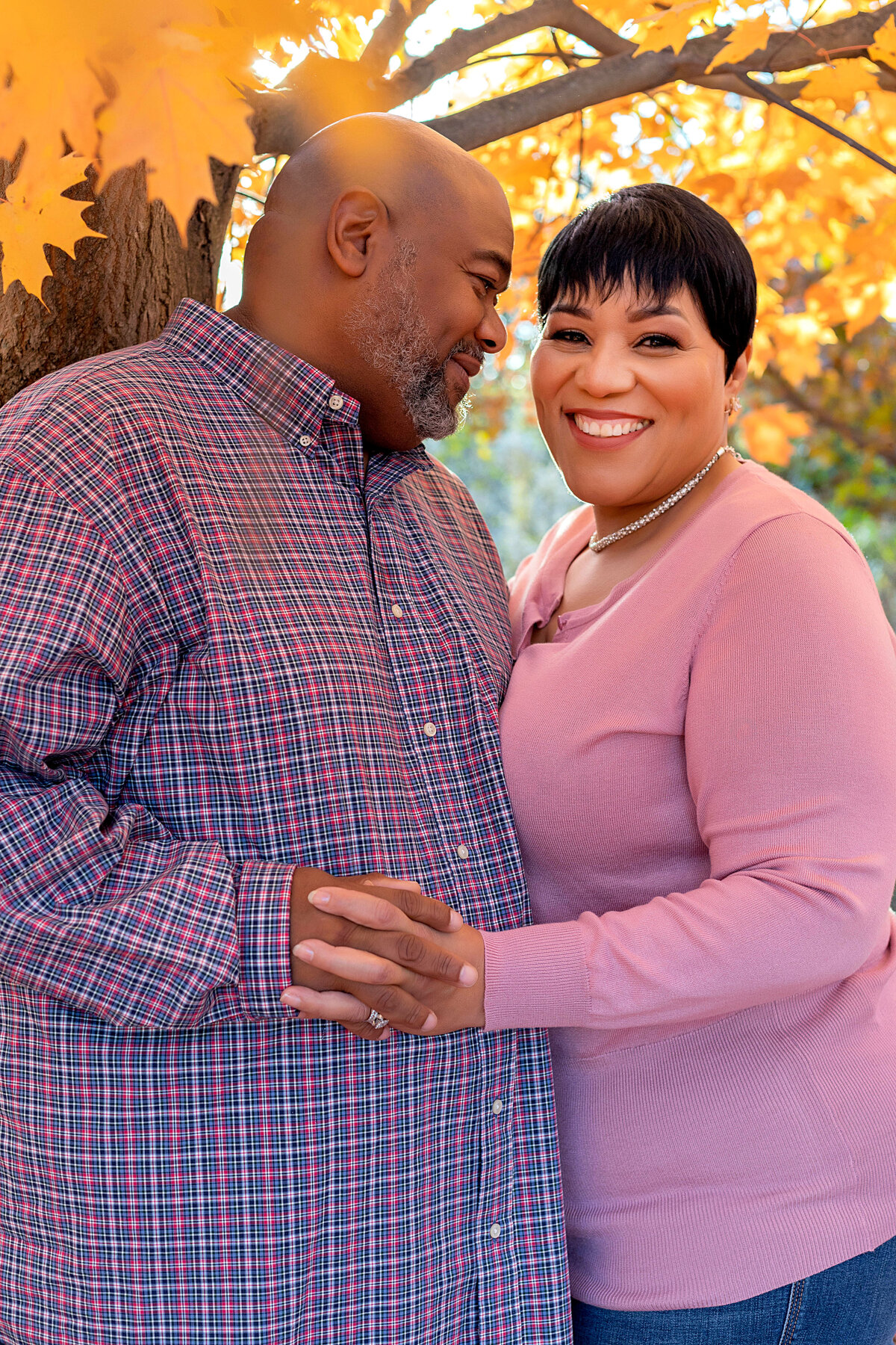 Married couple wearing blue and pink plaid top, pink sweater, and blue jeans standing under trees