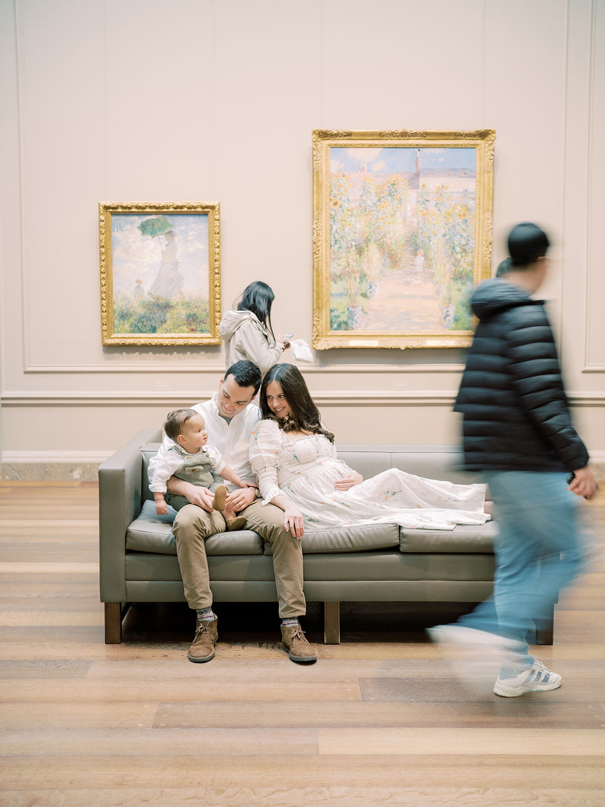 Mother and father sit on bench in National Gallery of Art with their toddler son as people walk by.