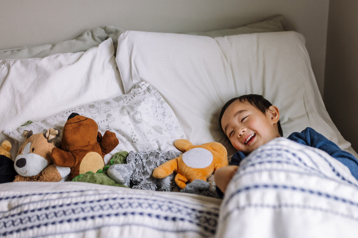 5-year-old boy laying in bed under covers smiling and looking at all of his stuffed animals also tucked under blanket