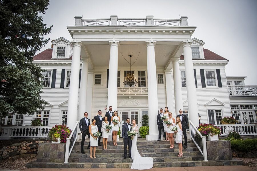 A bride and groom stand on the steps of The Manor House. Their wedding party stands along the steps on either side.