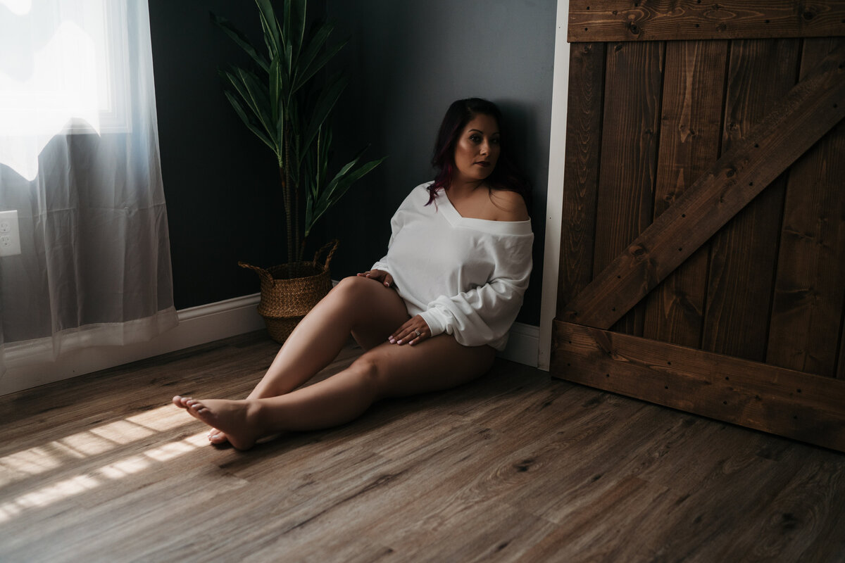 A woman in a white oversized shirt sits on the wood floor of a studio while leaning on a wall by a window and a barn door