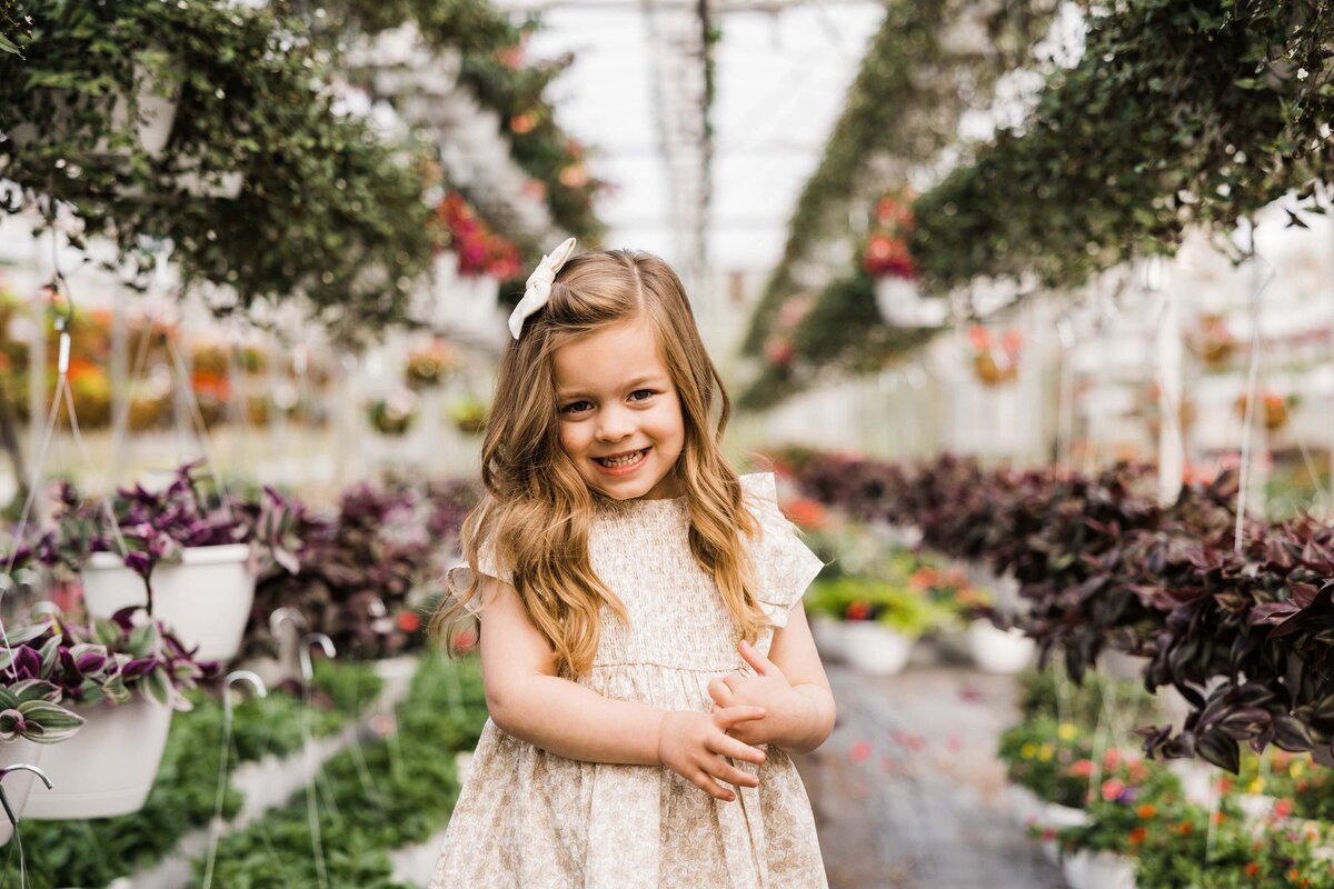 A smiling young girl standing in a greenhouse surrounded by hanging plants and flowers, captured by a family photographer Pittsburgh PA.