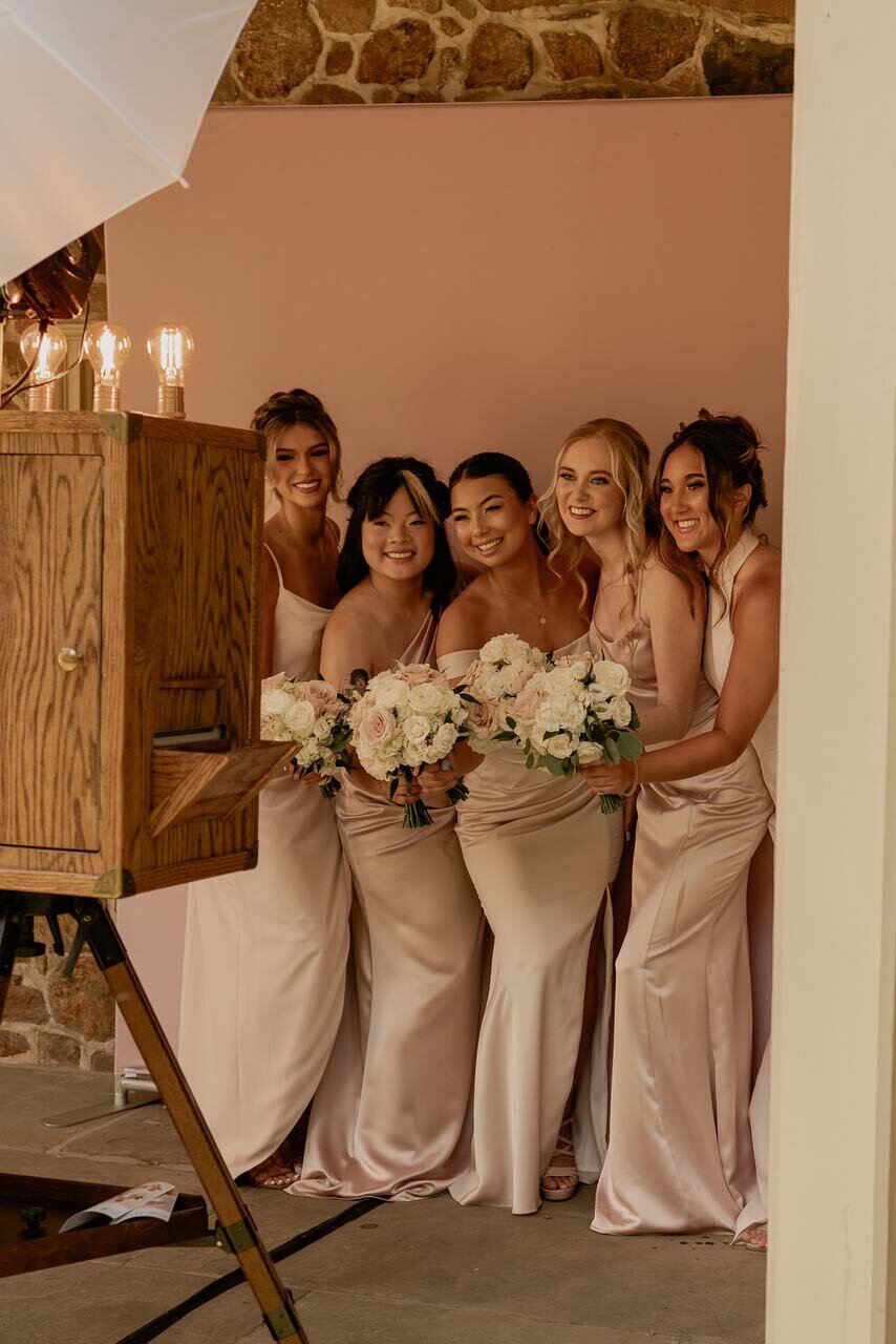 Bride in pink standing next to a vintage wooden photo booth under a chandelier with LOVE letters in the background.