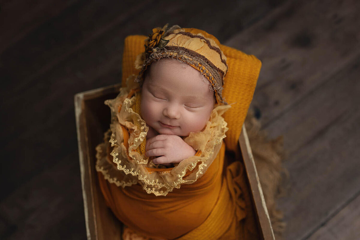 A newborn baby sleeps in a gold swaddle and matching bonnet in a wooden box