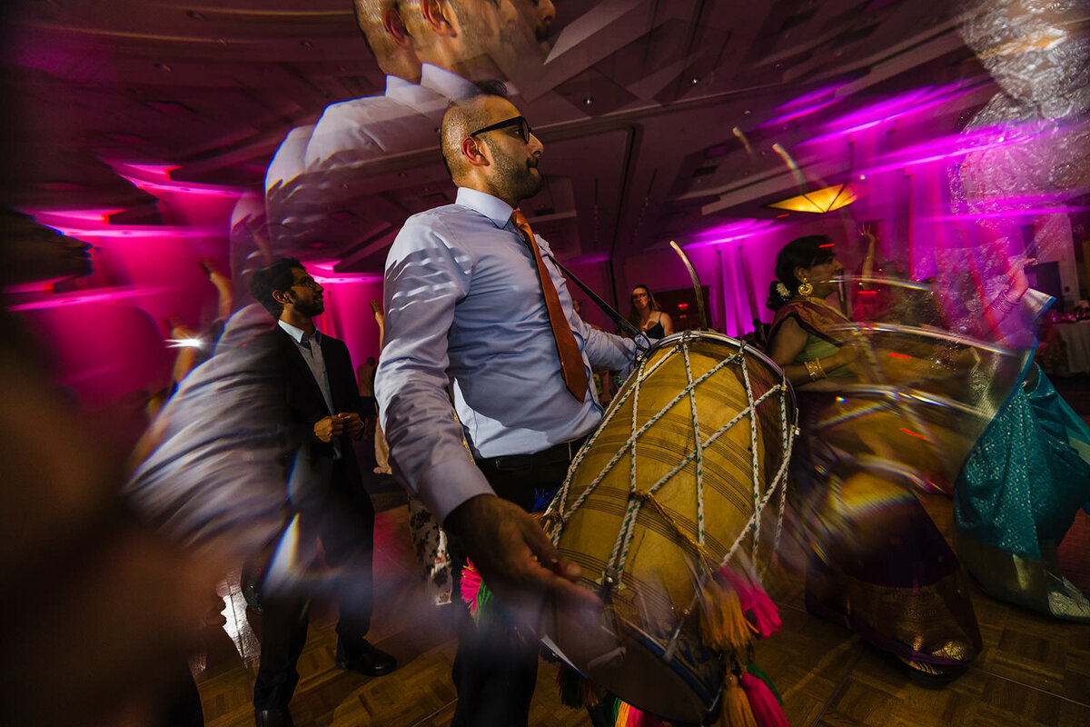 A drummer in motion plays a traditional dhol at a wedding event, with guests dancing around him in a vibrant, motion-blurred scene.