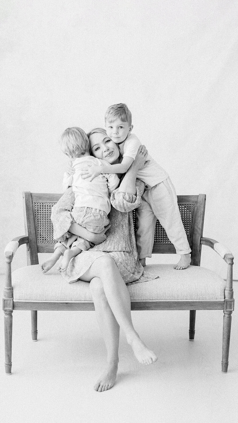 Black and white in studio photo of a mother sitting on a chair with her two boys snuggling her.