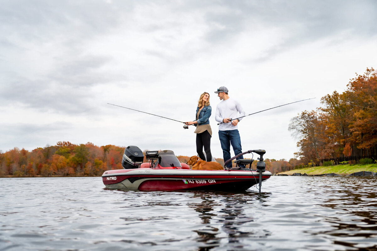 Man and woman, both standing on a fishing boat with fishing poles, smiling at each other, post for their engagement session in Mercer County Park