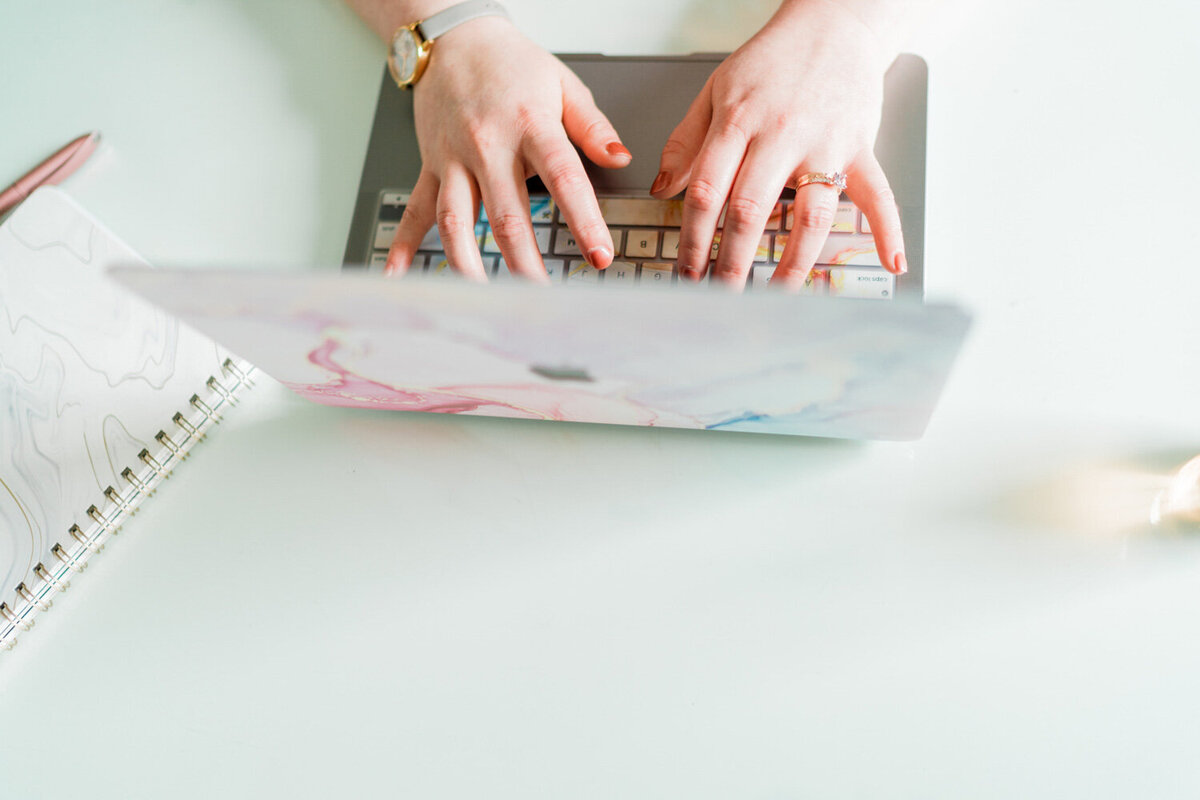 Becca Mai's hands typing on a colorful marble Mac lap top at a white desk next to a black and white marble spiral-bond notebook
