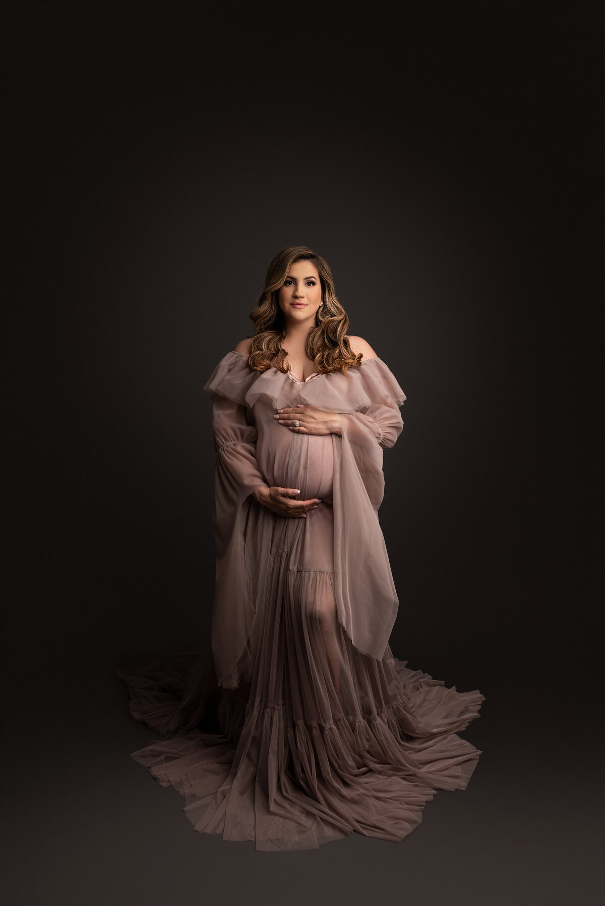 Maternity photo by Katie Marshall, Philadelphia Main Line's best maternity photographer. Woman in long organza blushy-taupe gown against dark grey backdrop, one hand above her bump, curled hair cascading over shoulders, closed-mouth smile, looking at the camera.