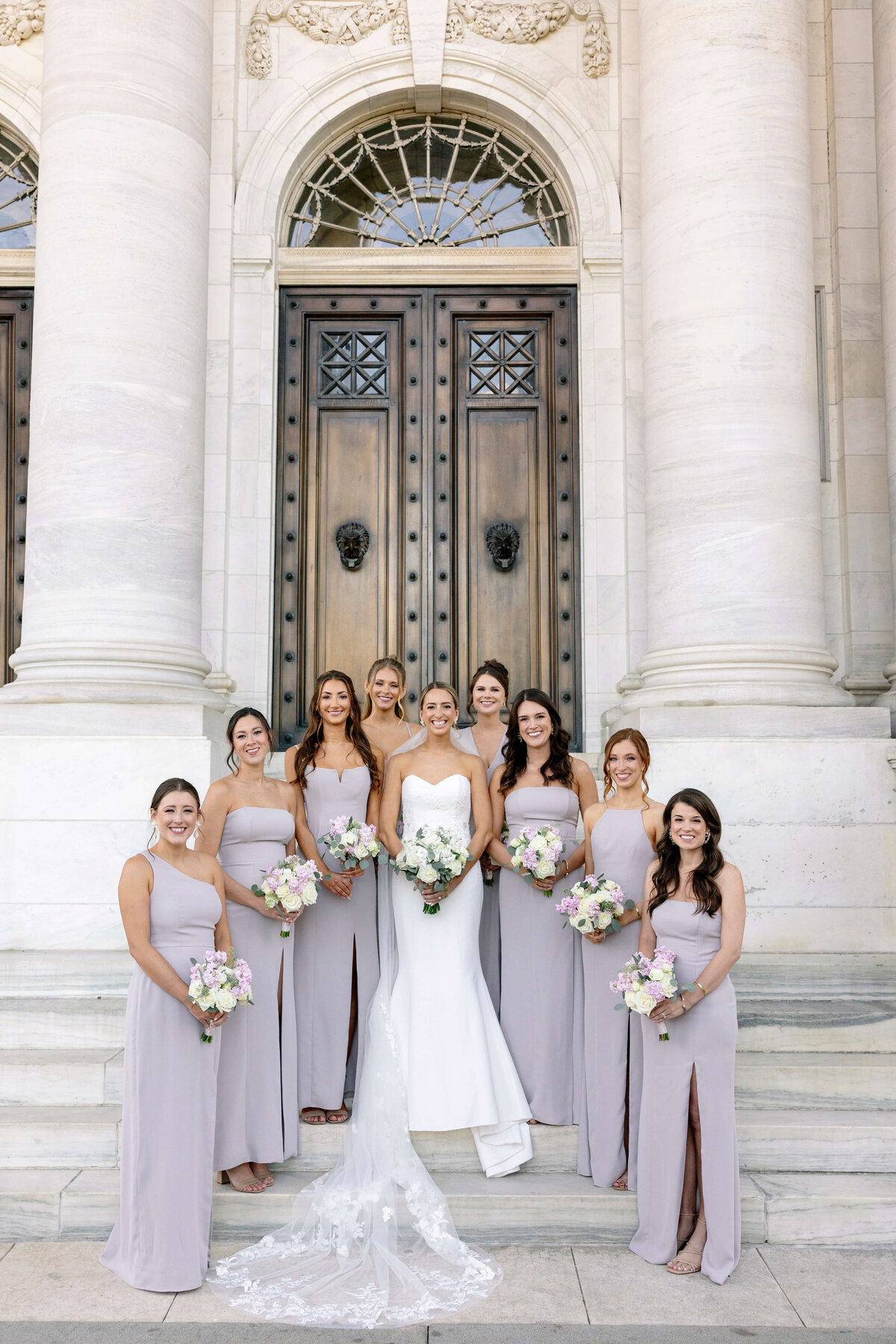 A bride stands on stone steps surrounded by eight bridesmaids outside a building with large wooden doors and columns. The bride wears a white gown, and the bridesmaids wear matching lavender dresses, each holding a bouquet.