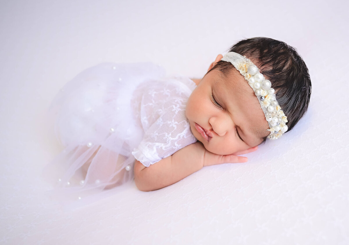 Newborn girl posed sleeping on her belly with a hand under her cheek.  She is in a white dress with a matching headband on a white blanket.  Image by Kitsap county photographer Beth Benecki.