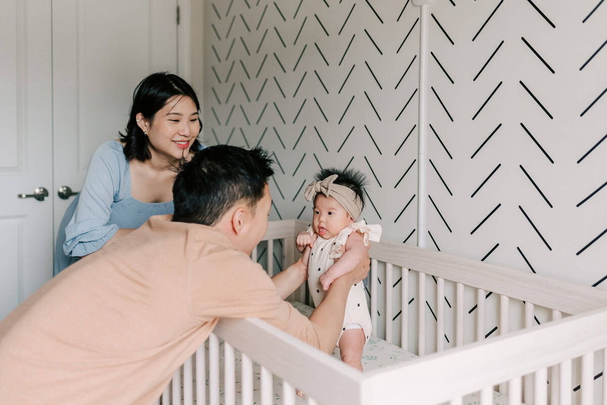 A dad holding his daughter up in her crib while mama looks on joyfully