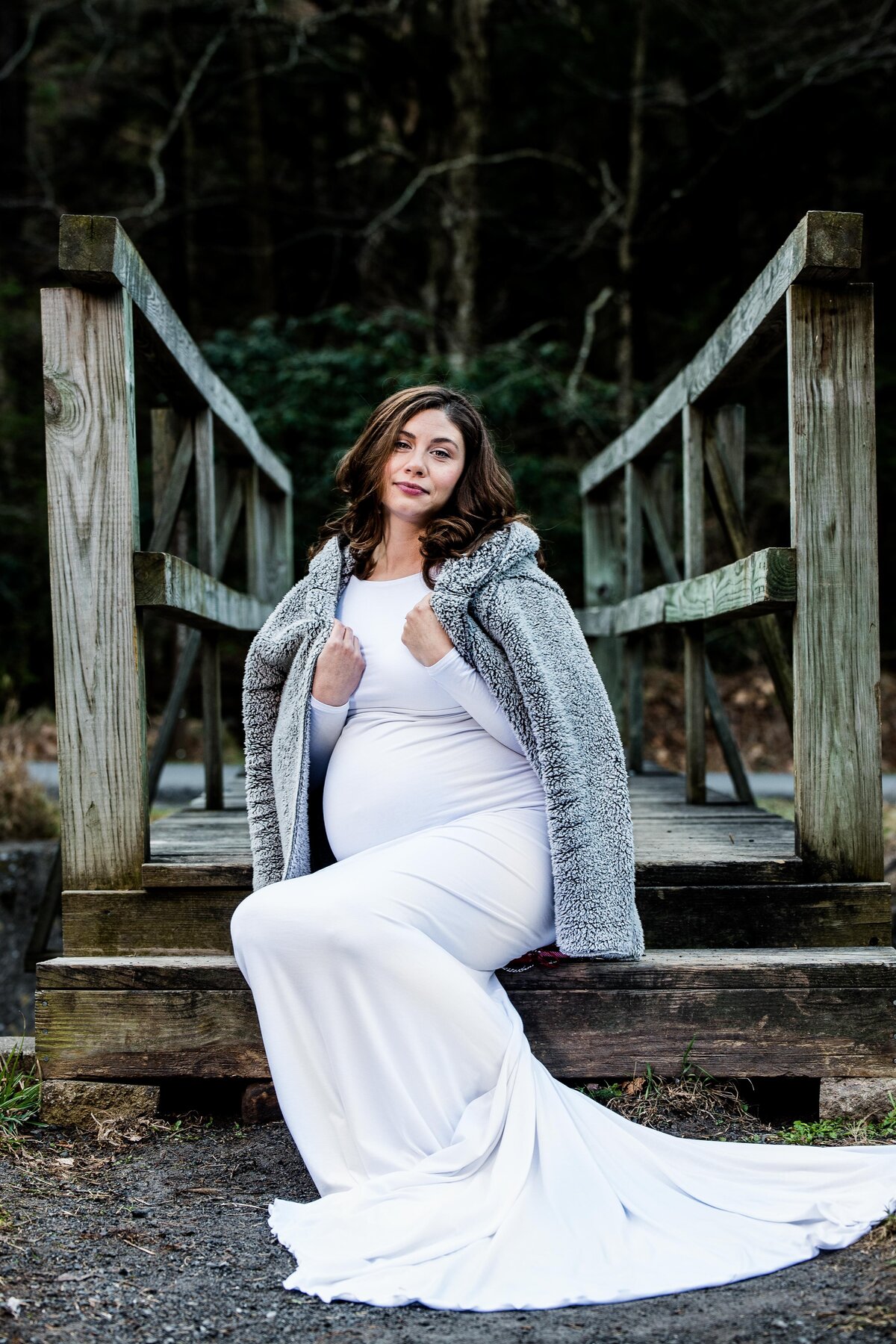 a pregnant woman wears a flowing white dress, holding a jean jacket over her shoulders
