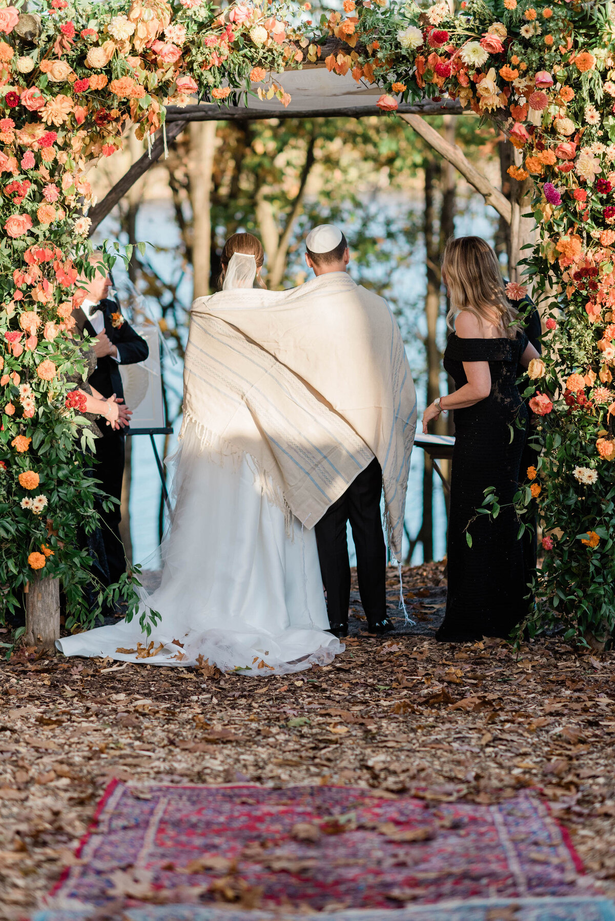 The couple stands under the chuppah with their backs to the camera, wrapped in a traditional Jewish tallit