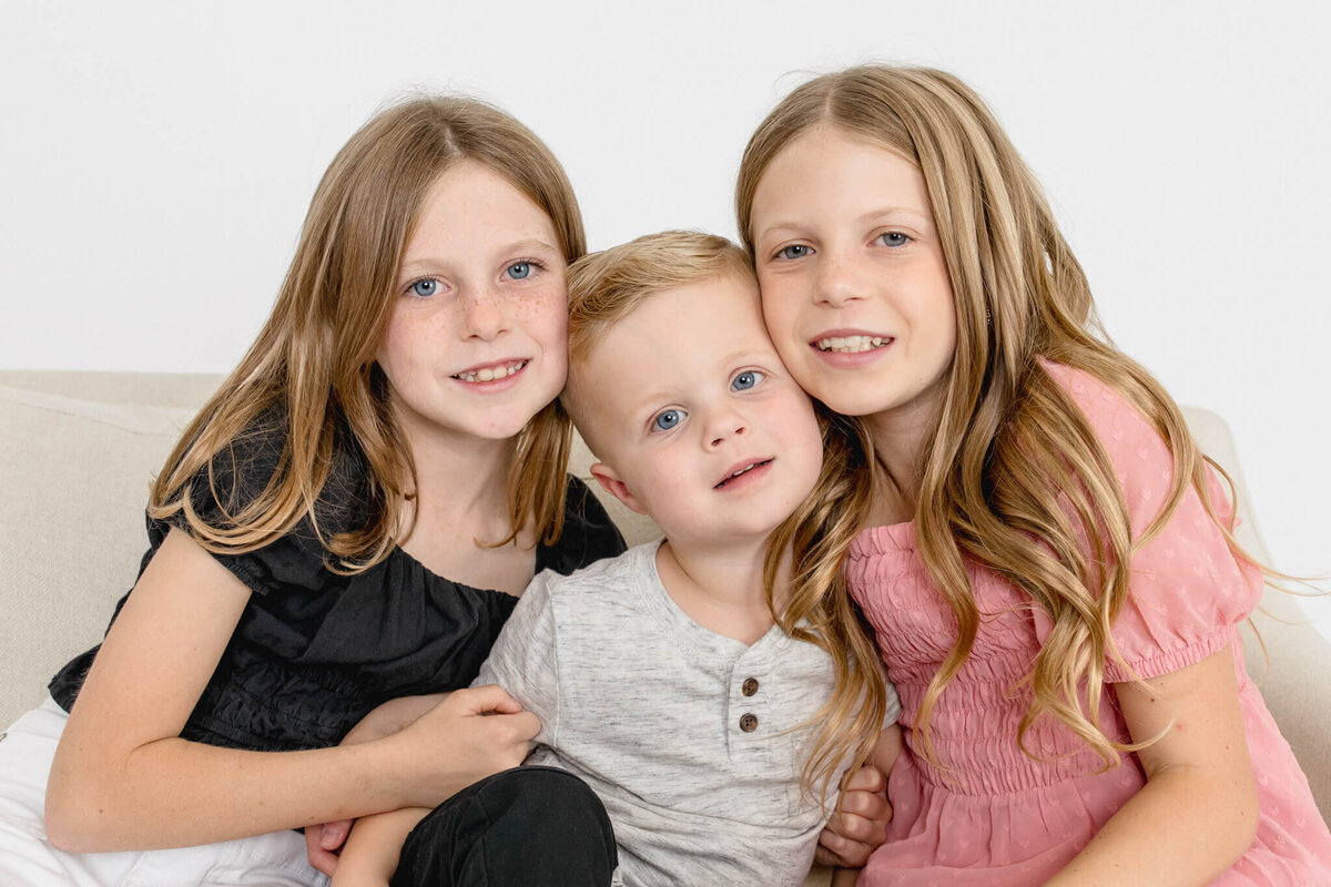 Two young girls putting their heads against their blonde little brother, all smiling at the camera in shades of black, pink, and grey in a white studio.