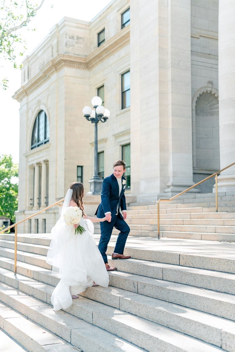A bride and groom walking up the grand steps of a majestic building, holding hands and smiling at each other.