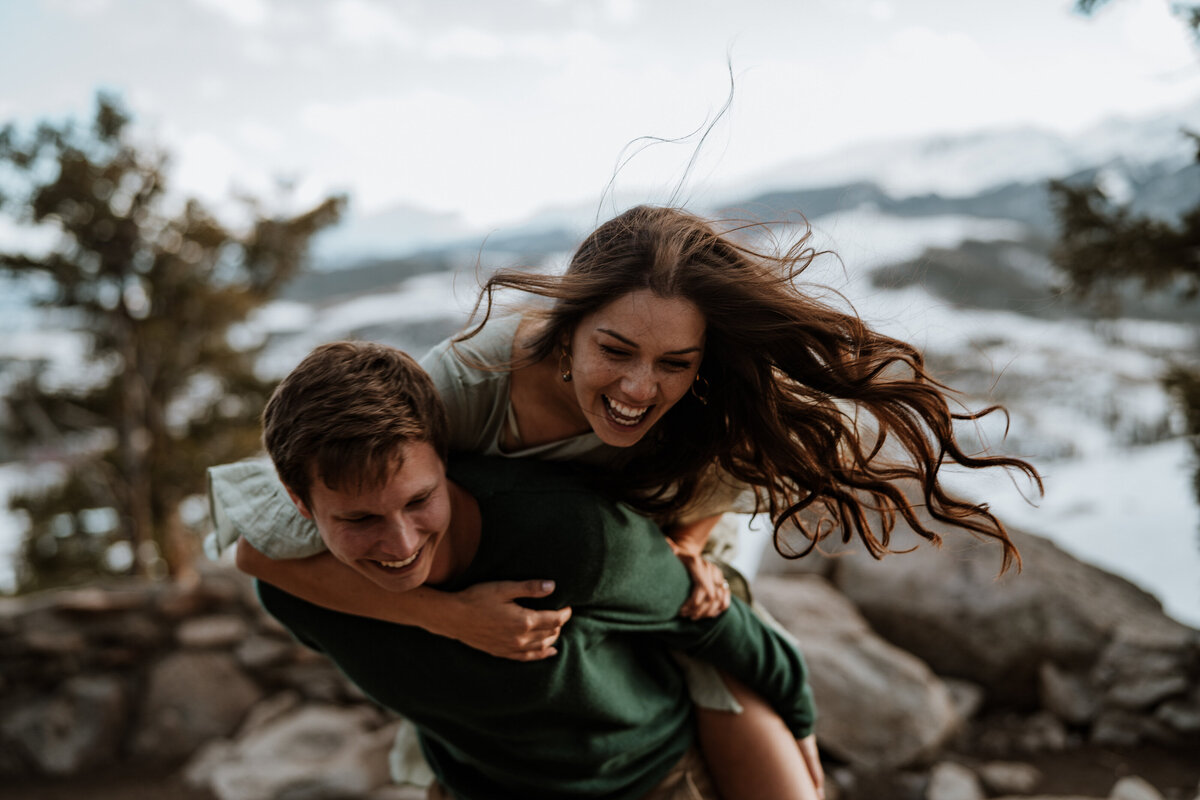 Couple laughs during playful piggyback ride with wind blowing her hair at Sapphire Point.