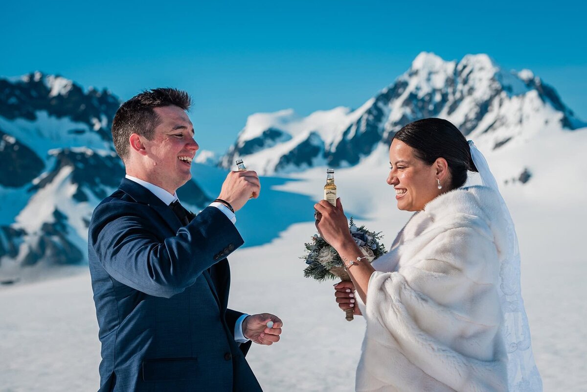 A bride and groom in their wedding finest laugh and smile and toast each other with tiny bottles of whiskey after just having exchanged elopement vows on the glacier during their two day adventure luxe Alaska elopement.   The sky is bright blue and the snow gleams white against the dark mountains rising up from the glacier.  