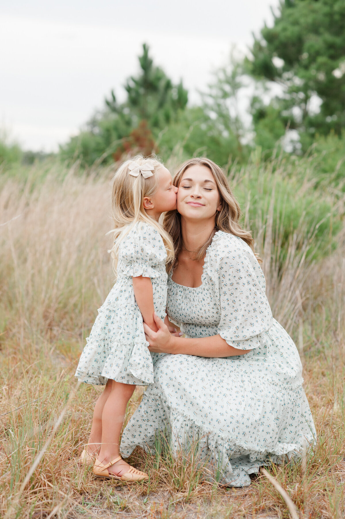Young daughter kissing moms cheek in a tall grass field at sunset