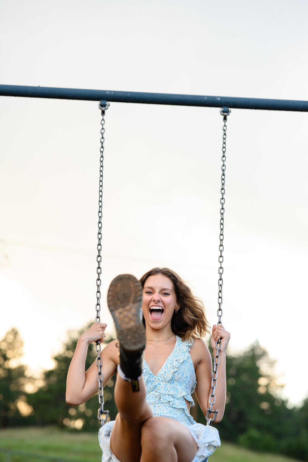 Fun senior pictures of a girl with the sun setting behind her while on a swing laughing with her foot extended