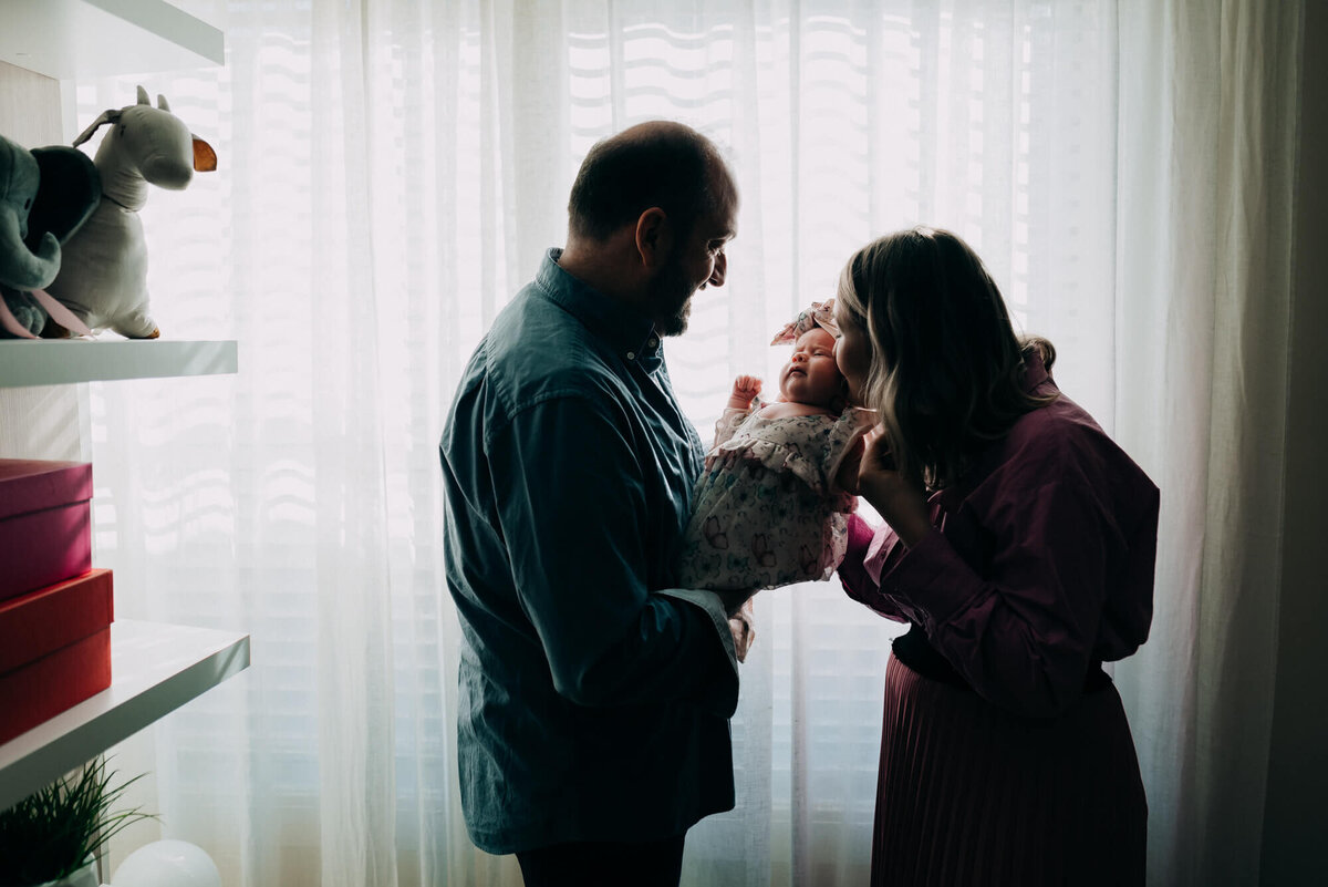 A mother and father hold their newborn daughter in front of their East Boston apartment window during their photoshoot with Allison Wolf, Boston Newborn Photographer