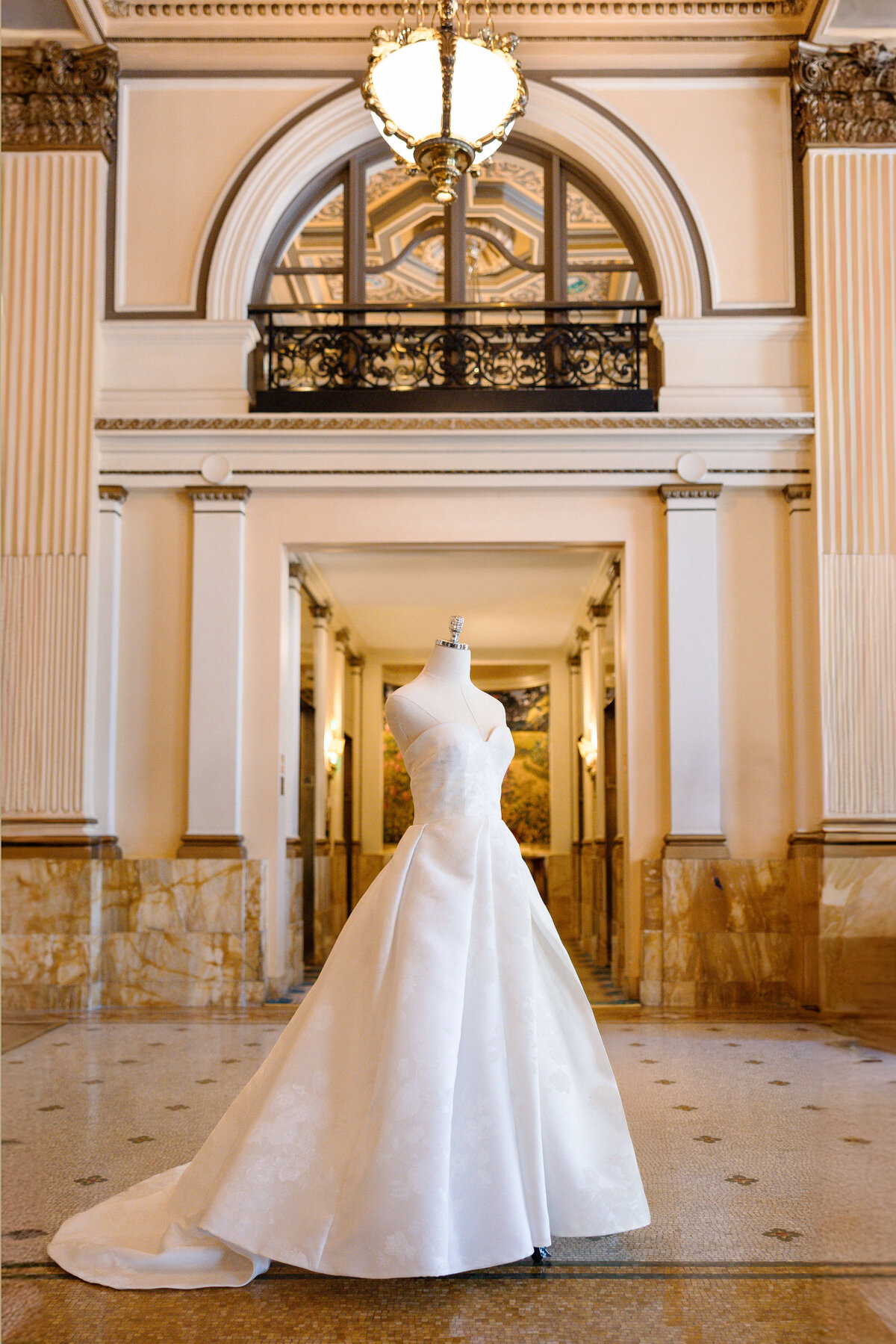 A luxurious white wedding gown displayed on a mannequin in an opulent interior, featuring tall marble columns, intricate molding, and a large chandelier overhead. The train of the dress elegantly drapes across the polished floor.