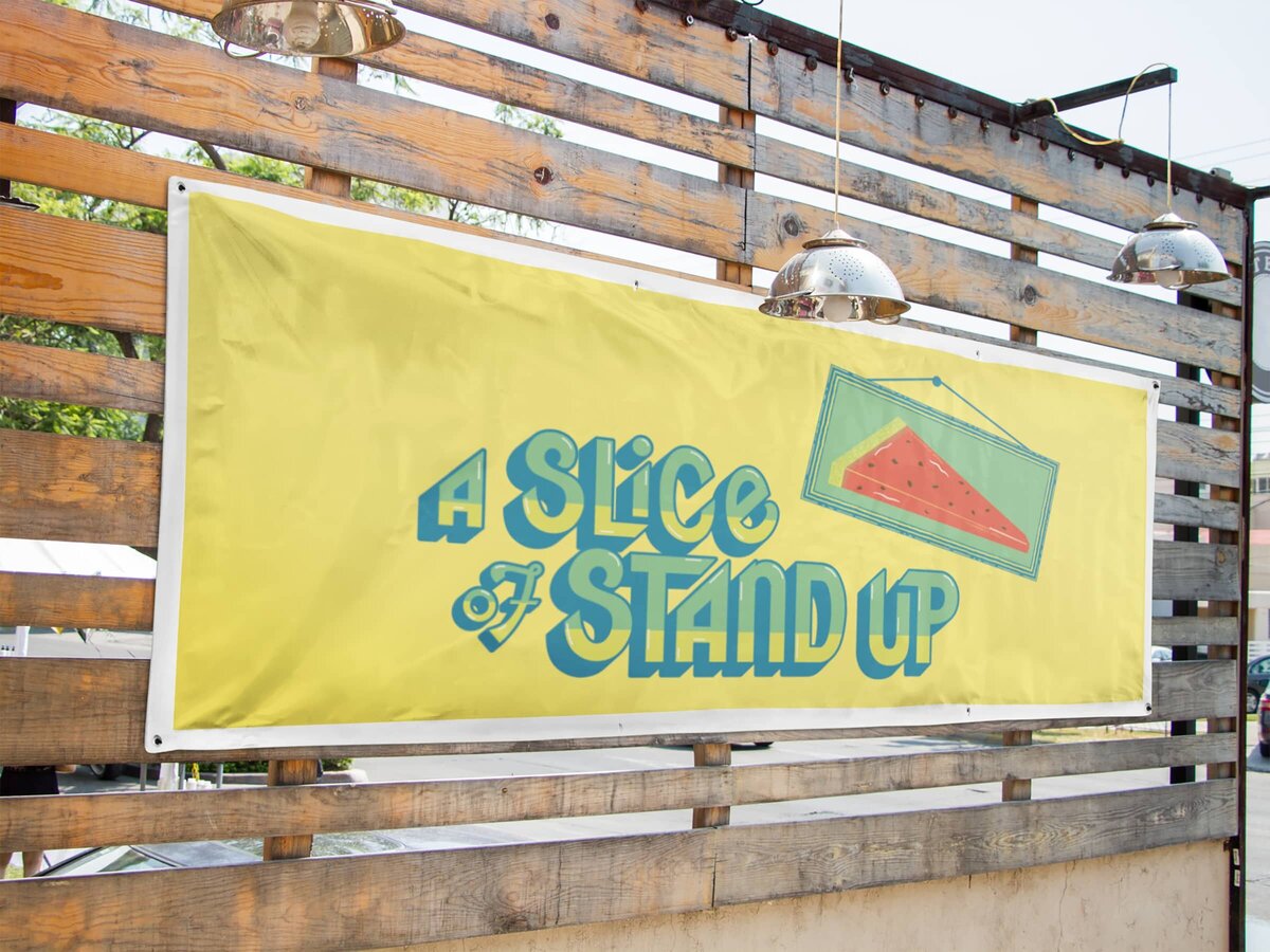 A Slice of Stand Up summer logo against a yellow background on a banner hanging against a wooden fence