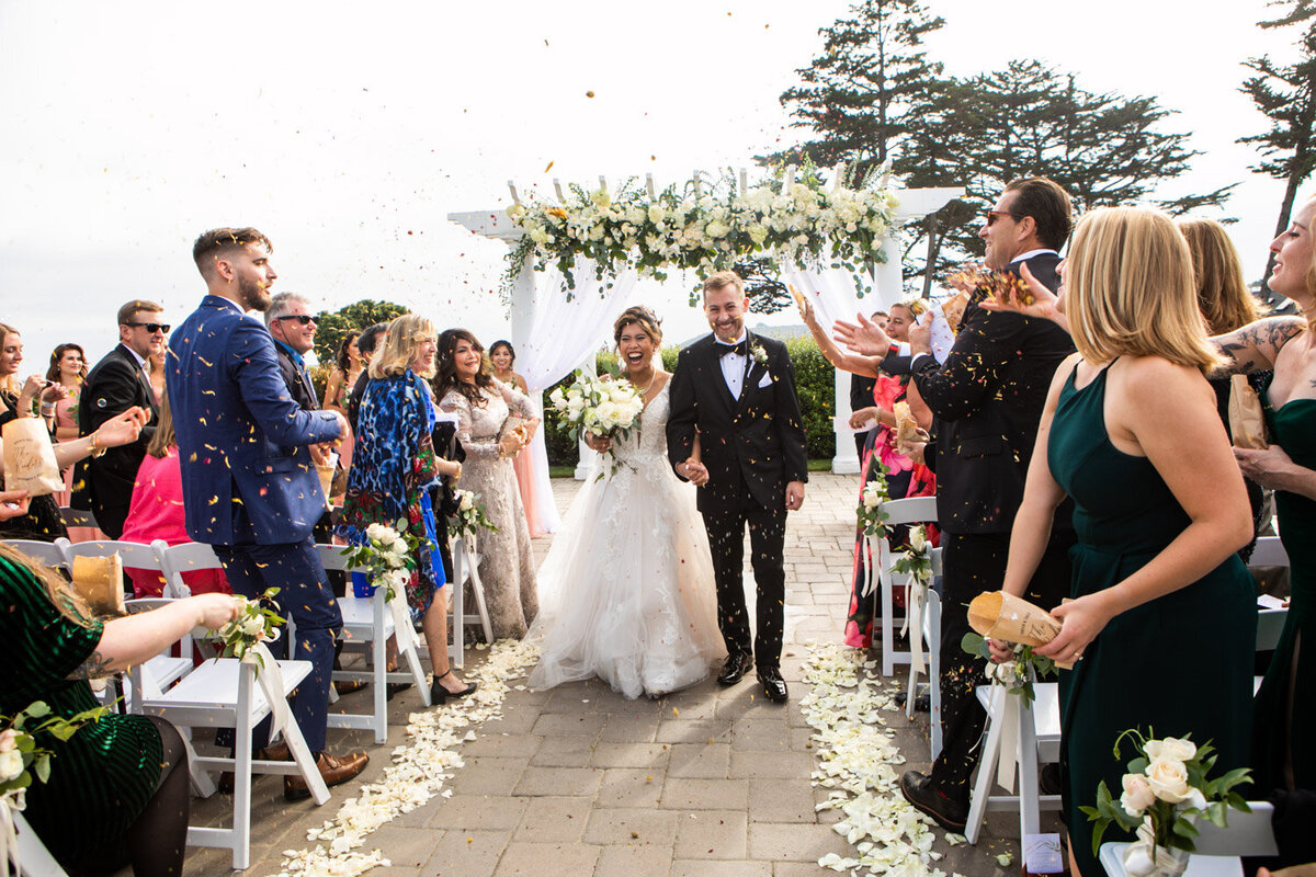 Wedding ceremony recessional under white floral arch with guests tossing petals
