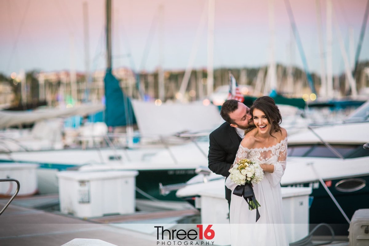 Bride leans forward smiling as the Groom kisses her cheek on the docks full of boats in Dana Point