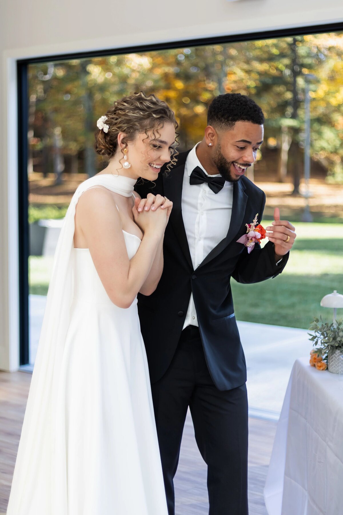 Wedding Photographers in RI | Kelsey Sheehan Photography Timeless Rhode Island Weddings | A joyful bride in a white dress and groom in a black tuxedo eagerly look at their wedding cake. They are indoors with a green park visible outside.