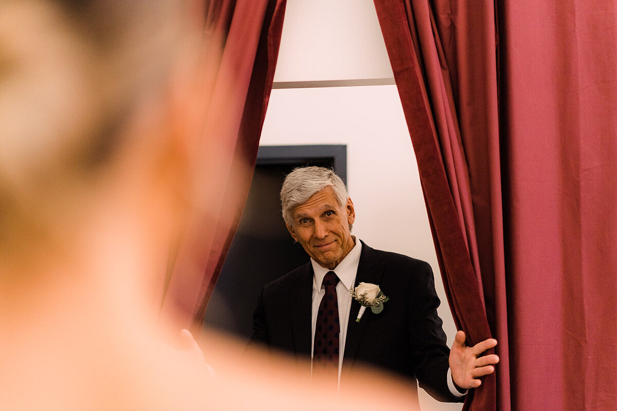 Father of the bride pulls back a curtain, smiling warmly as he catches a first glimpse of his daughter in her wedding dress.
