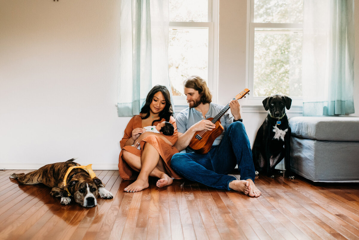 Newborn Photographer, Mom and dad sitting and playing the guitar for their baby while their dogs are around them.