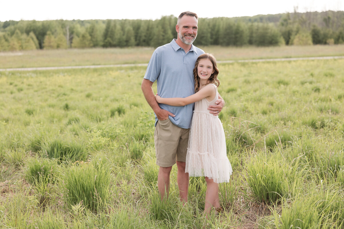 A father and his daughter are hugging in a field and smiling at the camera